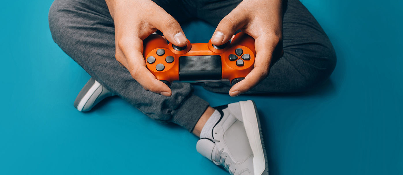 A young guy plays video games in his hands holding a red gamepad on a blue background, sitting in gray sneakers. Will esports ever rival traditional sport?