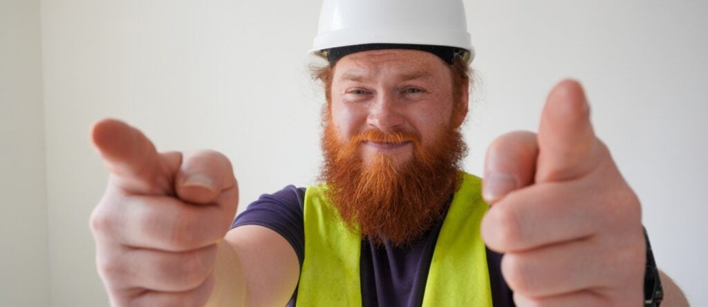 Bright smiling man in safety helmet and vest pointing at camera, construction or industrial worker.