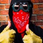 Person wearing a red bandana, goggles, and yellow gloves giving a thumbs-up against a brick wall.