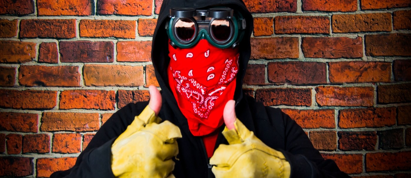Person wearing a red bandana, goggles, and yellow gloves giving a thumbs-up against a brick wall.